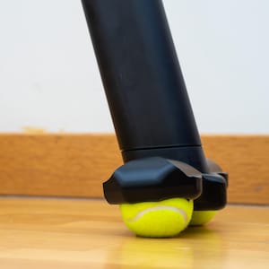 May include: A black furniture leg protector featuring a sleek, moulded design that securely holds two bright yellow tennis balls at the base, providing a cushioned contact point for the floor surface to prevent scratches and reduce noise.