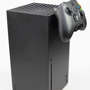 May include: A black Xbox Series X console with a black Xbox controller attached to the side. The console is standing upright on a white surface.