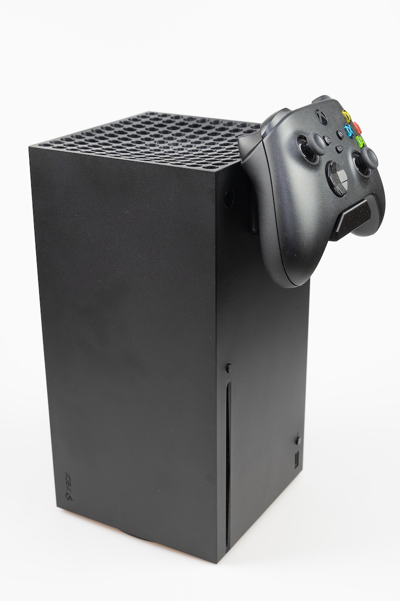 May include: A black Xbox Series X console with a black Xbox controller attached to the side. The console is standing upright on a white surface.