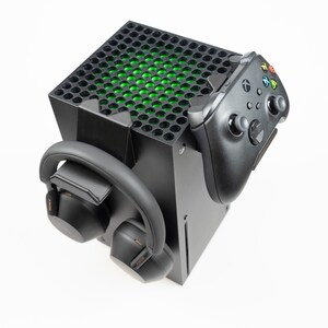 May include: A black gaming console stand with a black Xbox controller and black headphones attached to it. The console stand has a green light on the top.