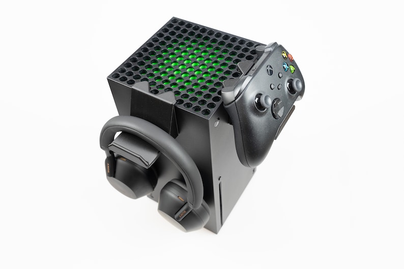 May include: A black gaming console stand with a black Xbox controller and black headphones attached to it. The console stand has a green light on the top.