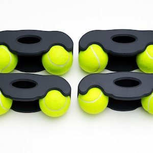 May include: Four black matte holders, each designed with a central oval cutout and two side openings, are each holding two bright neon yellow tennis balls. The holders are arranged in a two-by-two grid, showcasing a total of eight tennis balls.