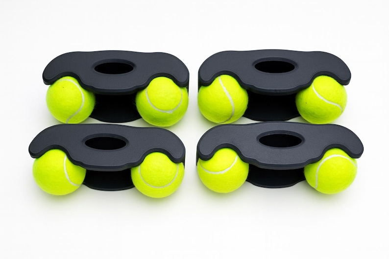 May include: Four black matte holders, each designed with a central oval cutout and two side openings, are each holding two bright neon yellow tennis balls. The holders are arranged in a two-by-two grid, showcasing a total of eight tennis balls.