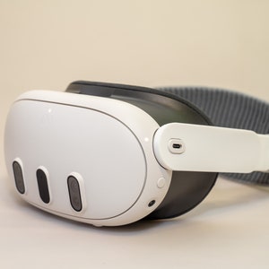 Może przedstawiać: Biały zestaw słuchawkowy do wirtualnej rzeczywistości z szarym paskiem z tkaniny. Zestaw słuchawkowy ma trzy czarne przyciski z przodu i port USB-C z boku.