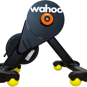 May include: A black Wahoo smart bike trainer featuring a circular flywheel with the white text wahoo and an orange center cap labeled Zwift. The base of the trainer is supported by four black legs, each equipped with a bright yellow tennis ball.
