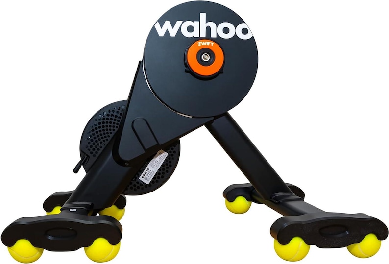 May include: A black Wahoo smart bike trainer featuring a circular flywheel with the white text wahoo and an orange center cap labeled Zwift. The base of the trainer is supported by four black legs, each equipped with a bright yellow tennis ball.