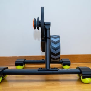 May include: A black bicycle trainer stand featuring a central vertical support, a circular flywheel with a grey chevron pattern, and four legs. Each leg base is fitted with a bright yellow tennis ball to provide stability and floor protection.