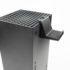 May include: A black gaming console with a stand attached to the top. The console has a black, perforated top with a green light visible through the holes.