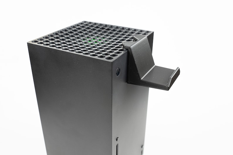 May include: A black gaming console with a stand attached to the top. The console has a black, perforated top with a green light visible through the holes.