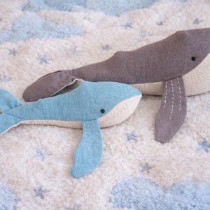 May include: Two plush whale toys. One is gray and the other is light blue. Both have white bellies and black eyes. They are lying on a white, fluffy blanket with blue cloud and star patterns.