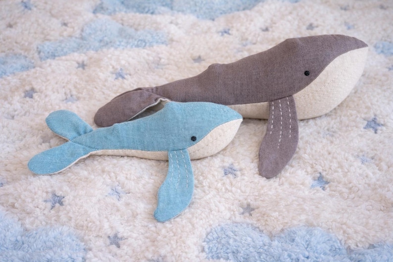 May include: Two plush whale toys. One is gray and the other is light blue. Both have white bellies and black eyes. They are lying on a white, fluffy blanket with blue cloud and star patterns.
