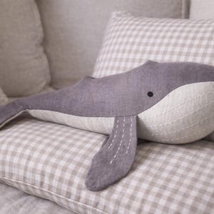 May include: A gray whale plush toy with a white belly and stitched details, resting on a checkered pillow. The whale has a small black eye and a curved body. The background features other pillows.