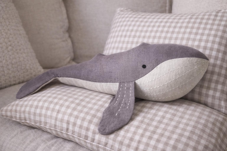May include: A gray whale plush toy with a white belly and stitched details, resting on a checkered pillow. The whale has a small black eye and a curved body. The background features other pillows.