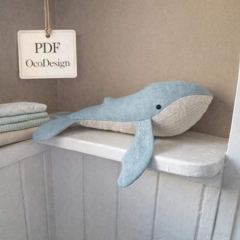May include: A light blue whale plush toy with a beige belly and fins, resting on a white shelf. The whale has a black eye and stitched details. A tag with "PDF OcoDesign" hangs nearby.