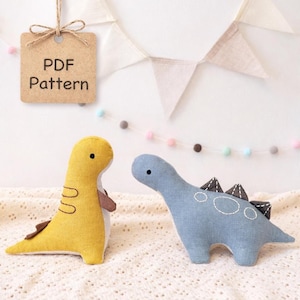 Puede incluir: Dos peluches de dinosaurio hechos a mano. Uno es amarillo con detalles marrones, el otro es azul con costuras blancas y púas grises oscuras. Una etiqueta dice "PDF Pattern". Los juguetes están sobre una superficie blanca con banderines decorativos.