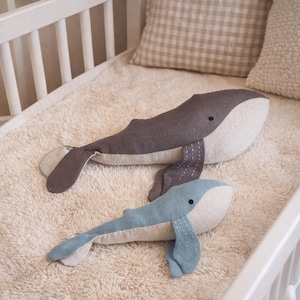 May include: Two plush whale-shaped stuffed animals in a crib. One is gray, the other is light blue, both with white bellies and stitched details. The crib has a white frame, and the bedding includes a fluffy cream-colored blanket and patterned pillows.