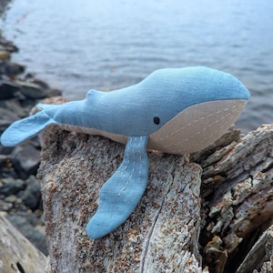 May include: A handmade blue whale plush toy with a beige belly and black eye. The whale is made of soft fabric and is resting on a rock near the water. The fins and tail are a lighter shade of blue.