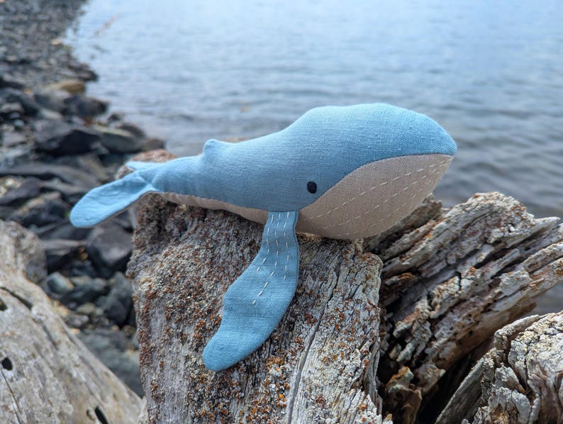 May include: A handmade blue whale plush toy with a beige belly and black eye. The whale is made of soft fabric and is resting on a rock near the water. The fins and tail are a lighter shade of blue.