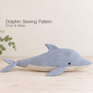 Op de afbeelding: Een stoffen dolfijn naaipatroon met een blauw en wit gestreepte bovenkant en een effen beige onderkant. De tekst Dolphin Sewing Pattern 31cm & 45cm is zichtbaar boven de dolfijn. De dolfijn heeft een klein zwart rond oog aan de zijkant.