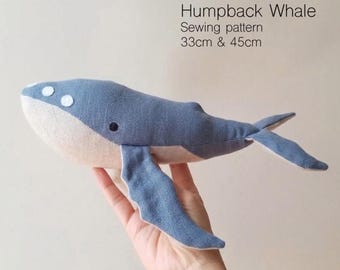 Op de afbeelding: Een naaipatroon voor een bultrug van blauwe en beige stof met witte cirkelvormige accenten op de kop en een donker oog. De tekst luidt Humpback Whale Sewing pattern 33cm & 45cm. Dit zachte object heeft een tweekleurig ontwerp met een blauwe bovenkant.