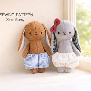 May include: Two plush bunny dolls, one brown and one gray, are displayed. The brown bunny wears blue and white striped shorts, while the gray bunny has a red bow and a white skirt. The text "SEWING PATTERN 20cm Bunny" is at the top.