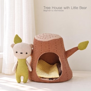 May include: A fabric tree house with a hollow center and a small stuffed bear toy. The tree house is brown with a light brown top and green leaf accents. The bear is light beige with a green body. Text reads: Tree House with Little Bear.
