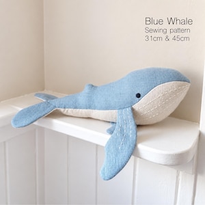May include: A blue whale sewing pattern plush toy with a light blue top and cream-colored underside. The whale features white stitched detailing on the fins and belly, a small dark eye, and text stating Blue Whale Sewing pattern 31cm & 45cm.