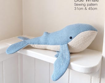 Op de afbeelding: Een blauwe walvis knuffel van een naaipatroon met een lichtblauwe bovenkant en een crèmekleurige onderkant. De walvis heeft witte stiksels op de vinnen en buik, een klein donker oog en de tekst Blue Whale Sewing pattern 31cm & 45cm.