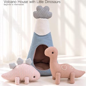 Puede incluir: Una casa volcán de tela en forma de cono con una parte superior gris de textura de gofre, cuerpo azul e interior marrón. Dos dinosaurios de peluche están delante: un estegosaurio rosa a rayas con placas grises y un dinosaurio rosa liso.