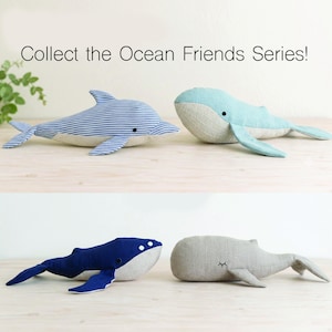 May include: Four plush sea animal toys from the Ocean Friends Series. One is a blue and white striped dolphin, one is a light blue whale, one is a dark blue whale, and one is a plain gray whale. All have simple stitched eyes and fins.