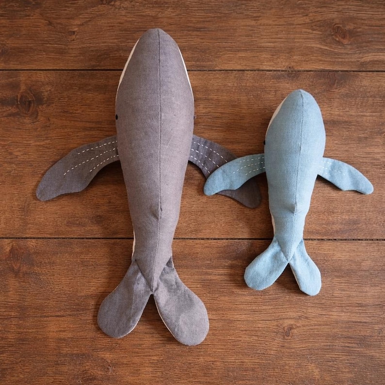 May include: Two handmade whale plush toys in shades of gray and blue, with white bellies and stitched details. The whales have outstretched fins and tail flukes, perfect for a nursery or child's room.