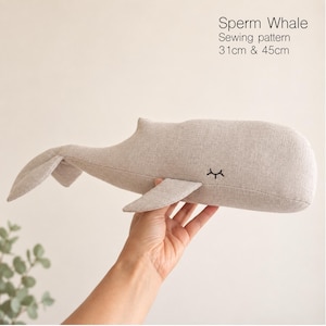 May include: A beige fabric sperm whale sewing pattern plush toy with embroidered closed eyes. The text on the image reads Sperm Whale Sewing pattern 31cm & 45cm. The whale features a rounded head, a single side fin, and a wide tail fluke.