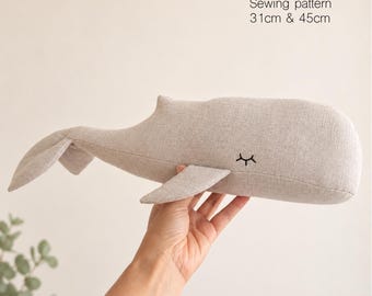 Op de afbeelding: Een beige stoffen potvis naaipatroon knuffel met geborduurde gesloten ogen. De tekst op de afbeelding luidt Sperm Whale Sewing pattern 31cm & 45cm. De walvis heeft een ronde kop, een zijvin en een brede staartvin.
