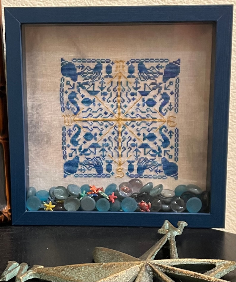 Op de afbeelding: Een ingelijst kruissteekontwerp met een nautisch thema in blauw en goud. Het ontwerp bevat zeepaardjes, ankers, vissen en andere nautische symbolen. De lijst is donkerblauw en het ontwerp is omringd door kleine blauwe glazen stenen.