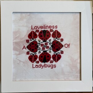 May include: A white framed cross-stitch with a red and black ladybug design. The text "A Loveliness Of Ladybugs" is stitched in red thread.