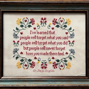 May include: A white cross-stitch embroidery framed in a dark wood frame with a teal blue mat. The embroidery features a quote by Dr. Maya Angelou: "I've learned that people will forget what you said, people will forget what you did, but people will never forget how you made them feel."