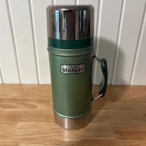 May include: A vintage Stanley thermos in a green and silver color scheme. The thermos has a handle and a lid. The Stanley logo is visible on the front. The thermos is cylindrical and appears to be made of metal.