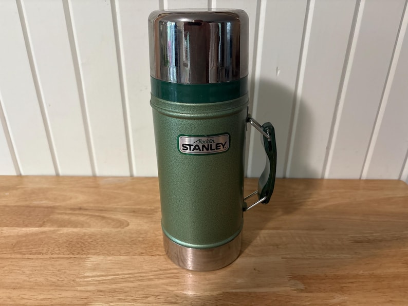 May include: A vintage Stanley thermos in a green and silver color scheme. The thermos has a handle and a lid. The Stanley logo is visible on the front. The thermos is cylindrical and appears to be made of metal.