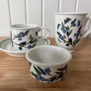 May include: A white ceramic teacup and saucer set, a mug, and a small bowl. Each item is adorned with blue flowers, green leaves, and butterflies. The teacup rests on a matching saucer with a green border. The mug and bowl also feature the floral design.
