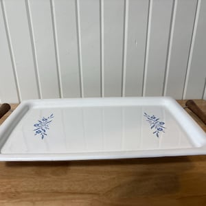 Könnte beinhalten: Weißes rechteckiges Serviertablett mit blauen Blumenmotiven und Holzgriffen. Das Tablett steht auf einer Holzoberfläche, mit einer weißen Wand im Hintergrund.
