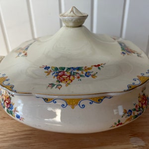 May include: Cream-colored, lidded serving dish with floral and gold trim. The dish features a rounded shape with handles and a decorative finial on the lid. Floral patterns include red, yellow, and blue flowers.