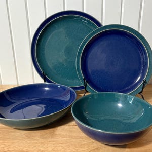Op de afbeelding: Een set van vier keramische gerechten in blauw- en tinten. De set bevat twee borden en twee kommen. De borden worden ondersteund door zwarte metalen standaards. De kommen zijn ovaal en rond. De gerechten hebben een gespikkelde textuur.