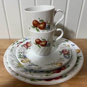 Pode incluir: Um conjunto de louça de cerâmica branca com duas canecas empilhadas. As canecas e os pratos apresentam um design de frutas com maçãs vermelhas, folhas verdes e bagas azuis. O conjunto inclui duas canecas e três pratos.