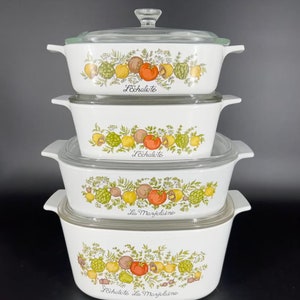May include: A set of four white ceramic casserole dishes with lids. The dishes are decorated with a floral pattern featuring green leaves, yellow lemons, red tomatoes, and brown mushrooms. The dishes are labeled "L'Echalote La Marjolaine".