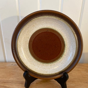 Può includere: Un piatto in ceramica con bordo marrone, fascia bianca e centro marrone rossiccio macchiettato. Il piatto è esposto su un supporto nero, mettendo in mostra il suo design. Questo piatto in stile vintage è adatto per servire cibo.