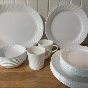 Könnte beinhalten: Ein Set aus weißem Geschirr mit einem wirbelnden Prägemuster an den Rändern. Die Kollektion umfasst eine große ovale Platte, einen runden Essteller, einen Stapel kleinerer runder Teller, einen Stapel kleiner Schüsseln und zwei passende Tassen.