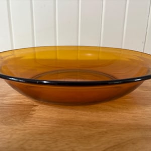 May include: A shallow, amber-coloured glass bowl. It has a wide design with a dark rim. The bowl is made of transparent glass and sits on a wooden surface.