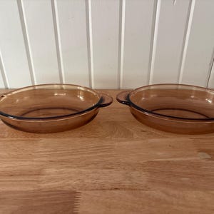 May include: Two amber-colored, oval-shaped glass baking dishes with handles. The dishes are empty and sit on a wooden surface. The background is a white wall with vertical wooden panels.