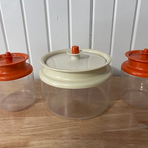 Op de afbeelding: Drie transparante plastic opbergcontainers met oranje deksels. De middelste container heeft een wit deksel.