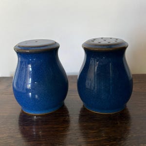 Pode incluir: Um par de saleiro e pimenteiro com um esmalte azul profundo brilhante e uma silhueta suave e arredondada. Cada peça apresenta uma borda castanho-escura na base e no topo. Um tem um único orifício na tampa, enquanto o outro tem vários.
